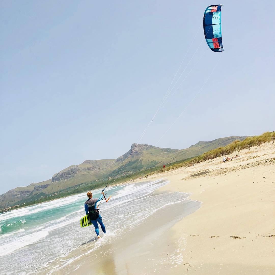 Johannes Herold walks along the beach at Son Serra de Marina with kite and board, the mountains of Mallorca behind him