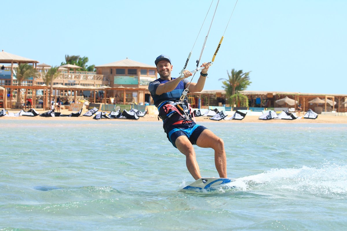 Johannes Herold wearing a cap, kitesurfing in shallow water in front of a beach with palm trees