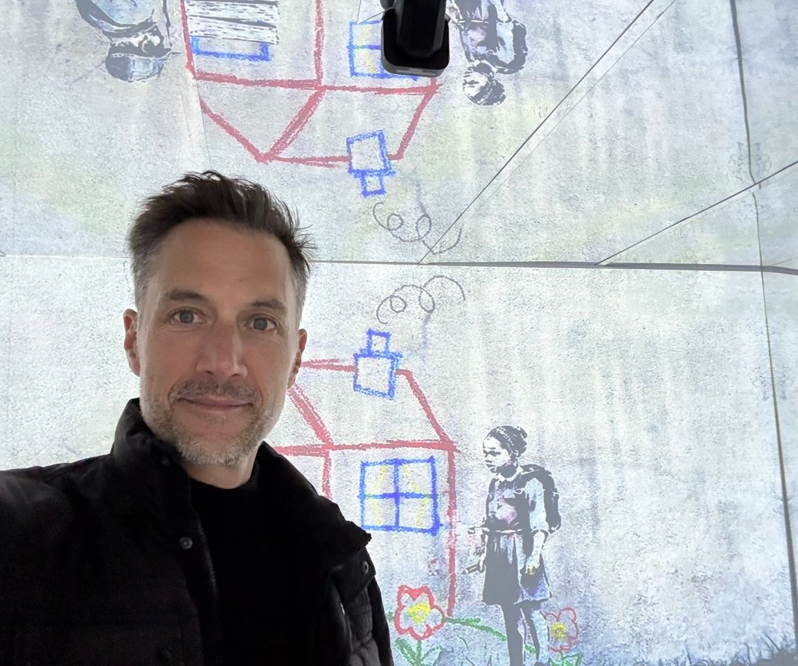 Peter Nötzel macht ein Selfie vor einer Wand mit Kreidezeichnungen von Häusern