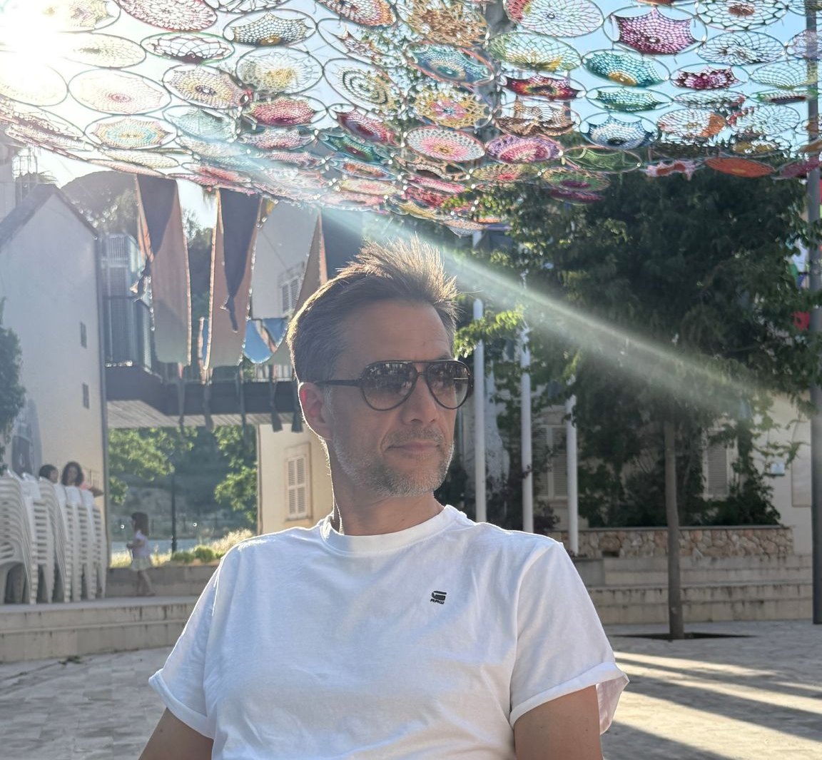 Peter Nötzel mit Sonnenbrille und weißem T-Shirt in der Sonne, über ihm bunte Schirme