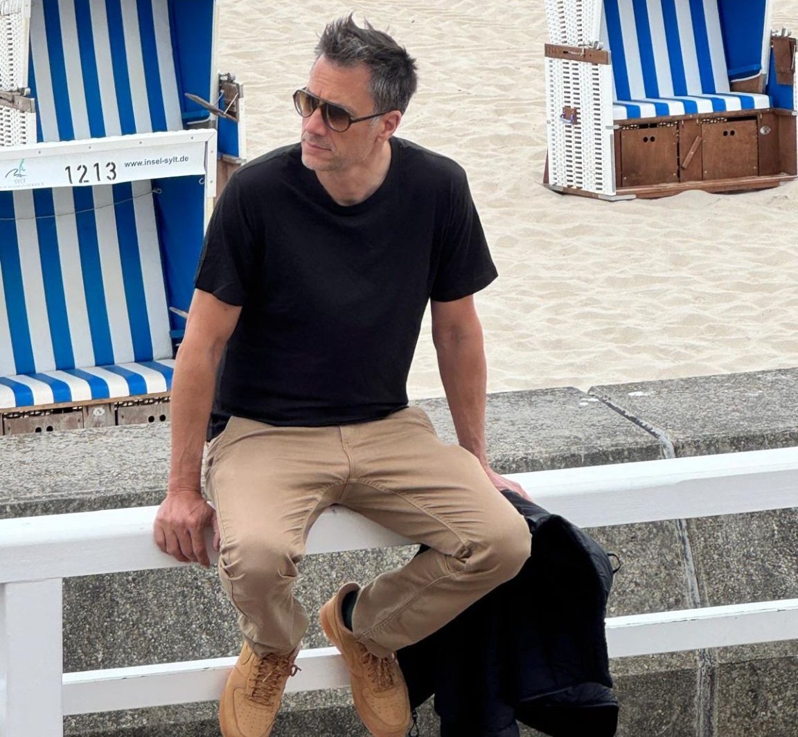 Peter Nötzel sitzt mit Sonnenbrille auf einer Bank vor blau-weißen Strandkörben auf Sylt
